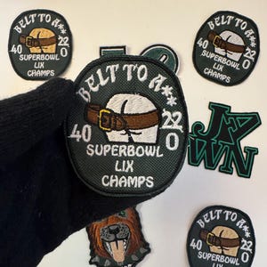 May include: A collection of embroidered patches with various designs. Several patches feature the text "BELT TO A**" and "SUPERBOWL LIX CHAMPS" with a graphic of a butt wearing a belt. Other patches include a dog and the letters "JVWN".