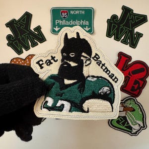 May include: A white embroidered patch with a cartoon of a person wearing a green and white football uniform and a black mask. The person has a beard and the text "Fat Batman" is written above and below the person.