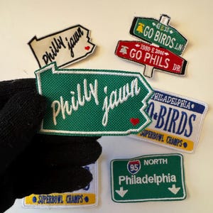 Könnte beinhalten: Eine Sammlung von bestickten Aufnähern mit Designs zum Thema Philadelphia. Die Aufnäher zeigen Sätze wie "Philly Jawn", "Go Birds" und "Go Phils" sowie ein Straßenschild für Philadelphia. Einige Aufnäher enthalten "Superbowl Champs".
