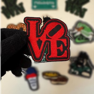 May include: A red and black embroidered patch with the word "LOVE" in bold letters. The patch is held by a black gloved hand, with other patches in the background, including a green Philadelphia sign and a green patch.