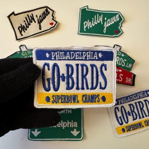 May include: Embroidered patches with Philadelphia-themed designs. One patch resembles a license plate with "GO-BIRDS" and "SUPERBOWL CHAMPS" text. Other patches say "Philly jawn" and street names. The patches are in various colors.