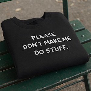 May include: Black sweatshirt with the phrase "PLEASE DON'T MAKE ME DO STUFF" printed in white. The sweatshirt is folded and resting on a green bench. The text is in a simple, sans-serif font.