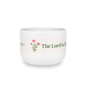 May include: A white ceramic mug featuring a horizontal band with a pink floral illustration and green text that reads The Lord is My and Psalm 28:7. The mug has a rounded, wide-rimmed shape with a smooth, glossy finish and a clean, simple design.