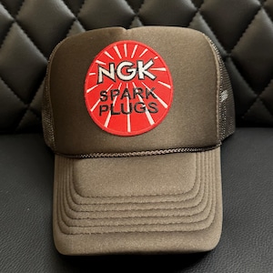 May include: A brown trucker hat with a red and white embroidered patch that says "NGK SPARK PLUGS".