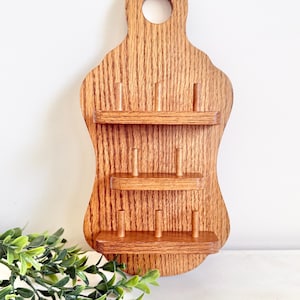 May include: A wooden wall-mounted thread rack with three shelves and multiple holes for storing thread spools. The rack is stained a rich brown color and has a decorative cutout at the top.