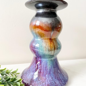 May include: A handcrafted ceramic candle holder with a unique, bulbous shape. The color gradient transitions from a glossy black top to shades of orange, teal, and purple. The base has a speckled, textured finish, adding visual interest to this decorative piece.
