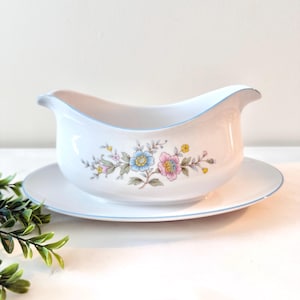May include: White ceramic gravy boat with a matching saucer. The gravy boat features a floral design with blue, pink, and yellow flowers. A light blue trim accents the edges of both pieces. The set is designed for serving sauces and gravies.