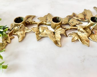 May include: A gold-colored metal candle holder with three circular openings for candles. The candle holder is shaped like a vine with detailed leaf designs. The leaves and vine are arranged horizontally.