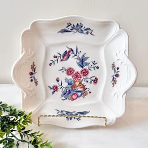 May include: White square serving platter with a scalloped edge. The platter features a floral design with pink, blue, and red flowers and leaves. The platter is displayed on a gold-colored metal stand.