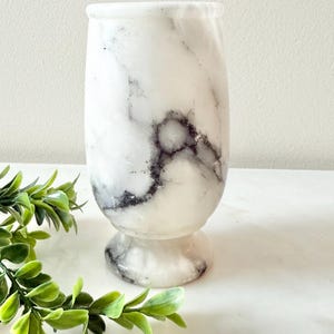 May include: A white marble vase with gray veining. The vase has a rounded base and a cylindrical body with a slightly flared rim. The marble has a natural, organic pattern, and the vase is set against a neutral background.