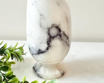 May include: A white marble vase with gray veining. The vase has a rounded base and a cylindrical body with a slightly flared rim. The marble has a natural, organic pattern, and the vase is set against a neutral background.