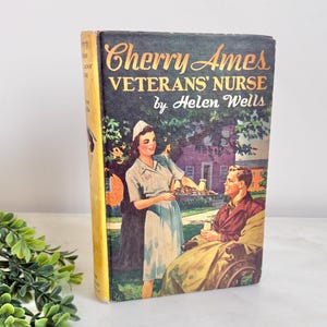 May include: Vintage hardcover book titled "Cherry Ames Veterans' Nurse" by Helen Wells. The cover features an illustration of a nurse serving a tray to a man in a wheelchair. The book has a yellow spine and a dark green background.