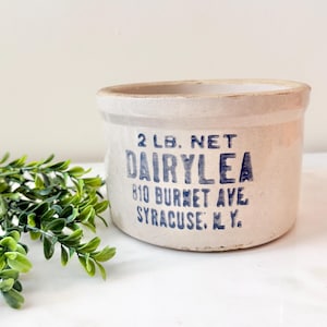 May include: A cylindrical off-white stoneware crock with a blue printed label that reads 2 LB. NET DAIRYLEA 810 BURNET AVE SYRACUSE, N.Y. The container features a thick rim and a smooth, matte finish, presenting a classic utilitarian design.