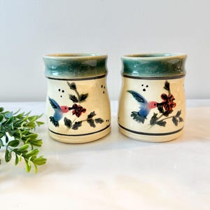 May include: Two ceramic jars with a cream-colored base, adorned with hand-painted hummingbirds and flowers. The jars have a green rim and a black base. The hummingbird's wings are blue, and the flowers are red.