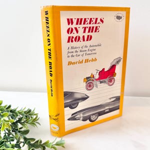May include: A vintage book titled "WHEELS ON THE ROAD" by David Hebb. The cover is yellow with red and black text, and features illustrations of classic cars. The book's spine also displays the title. The book is a history of the automobile.