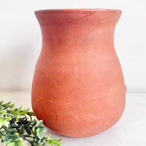 May include: A terracotta-colored vase with a wide base and a narrow neck. The handmade pottery has a textured surface and a rustic appearance. The vase is approximately 10 inches tall and is suitable for displaying flowers or as a decorative piece.