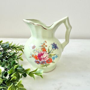 May include: A light green ceramic pitcher with a floral design. The pitcher features a handle with a unique, textured design. The floral pattern includes pink, red, orange, blue, and purple flowers. The rim has a distressed, brown edge.