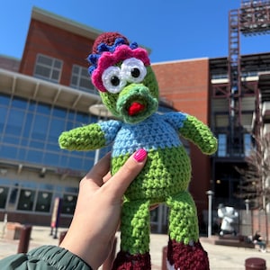 May include: A handmade crochet doll with a whimsical design. The doll features a green body, blue and green arms, and burgundy and white shoes. It has a blue and pink hat and large white eyes. The doll is held up in front of a building.