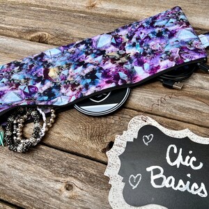 May include: A purple and blue zippered pouch with a geometric pattern. The pouch is open and filled with various items, including a black cord, a silver bracelet, and a black disc. The pouch is sitting on a wooden surface with a sign that reads "Chic Basics".