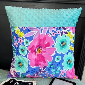 May include: A turquoise blue pillow with a pocket on the front. The pocket is lined with a floral fabric featuring pink, purple, and green flowers on a blue background. The pillow is on a bed with a light blue blanket.