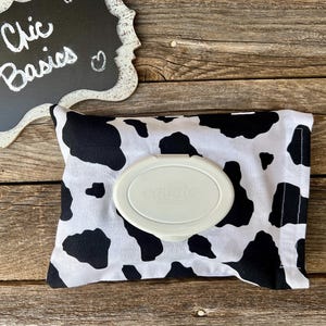 May include: A black and white cow print fabric wipe dispenser with a white plastic wipe container. The dispenser has a black border and the text "Chic Basics" is written on a chalkboard in the background.