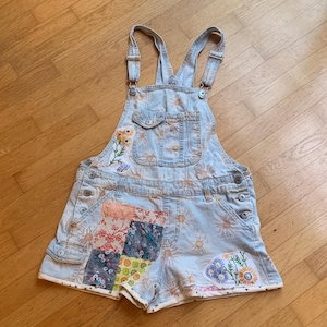 May include: Light blue denim overall shorts featuring an all-over daisy print, adjustable shoulder straps, and a front bib pocket. The garment includes a patchwork panel on the left leg and embroidered floral accents on the bib and right leg.