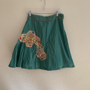 Pu&ograve; includere: Gonna color verde acqua con un design patchwork decorativo. La gonna presenta una serie di toppe esagonali in vari colori e motivi, aggiungendo un tocco unico. La gonna &egrave; appesa a una gruccia contro un muro bianco.