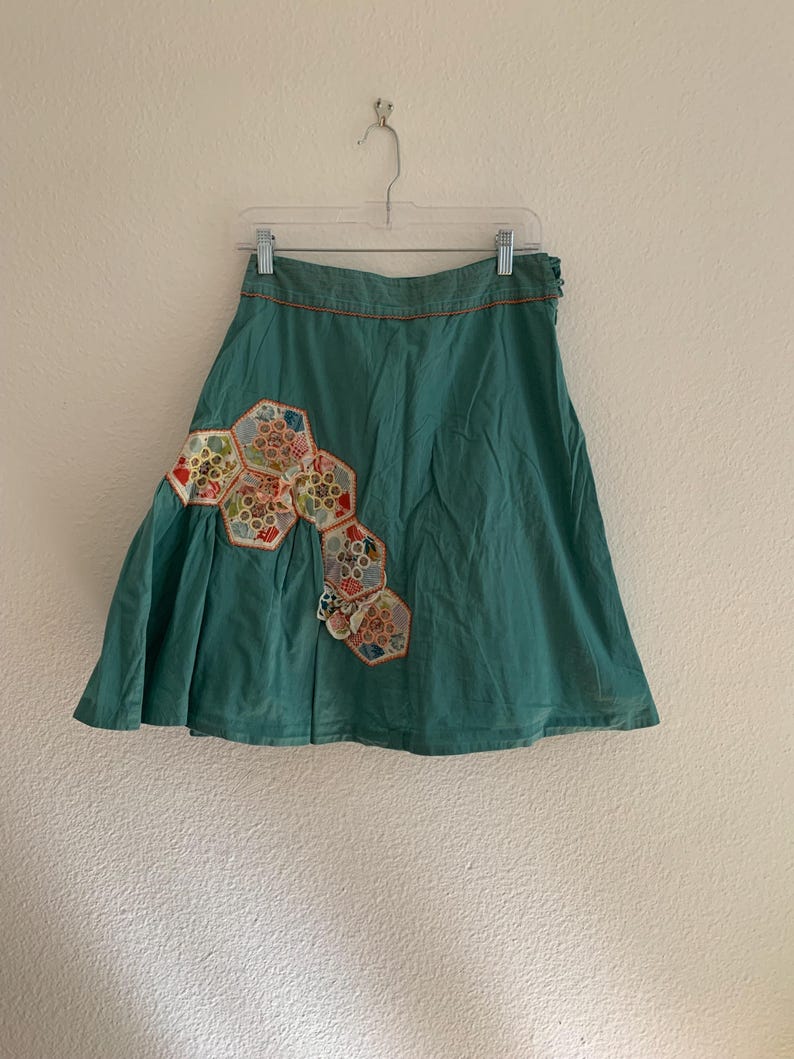 Pu&ograve; includere: Gonna color verde acqua con un design patchwork decorativo. La gonna presenta una serie di toppe esagonali in vari colori e motivi, aggiungendo un tocco unico. La gonna &egrave; appesa a una gruccia contro un muro bianco.