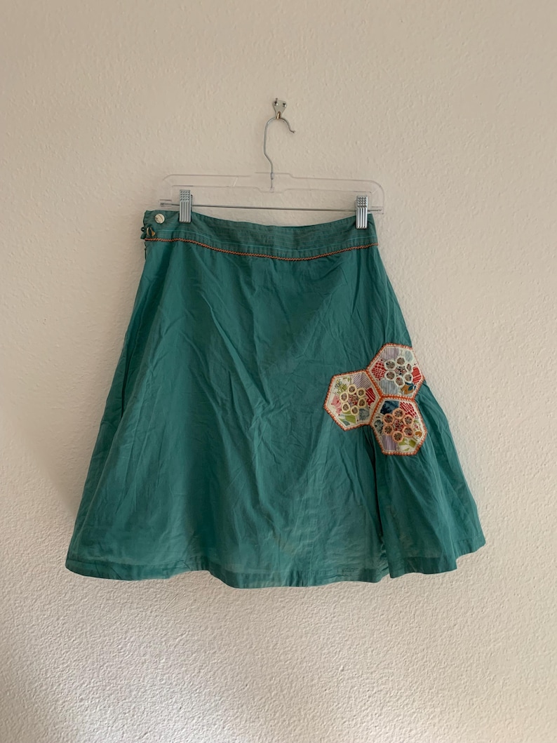 Pu&ograve; includere: Una gonna color teal con una silhouette svasata a trapezio. La gonna presenta una toppa decorativa di forme geometriche colorate in tessuto sul davanti. &Egrave; appesa a una gruccia trasparente contro un muro bianco.