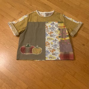 May include: A patchwork short-sleeved shirt with a variety of patterns and colours. The shirt features a combination of floral, striped, and solid fabric panels. Two embroidered pumpkins are on the front. The collar and sleeve cuffs are trimmed with a contrasting fabric.