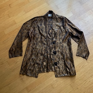 May include: A long-sleeved, button-down jacket in a brown and gold floral pattern. The jacket has three black buttons and a collar. The fabric has a shiny, textured appearance. The jacket is laid flat on a wooden surface.