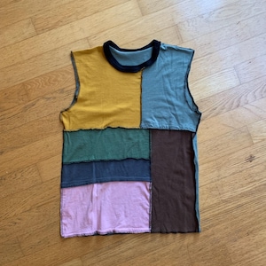 以下が含まれることがあります： パッチワークデザインのノースリーブトップス。マスタードイエロー、ティール、グリーン、ネイビー、ピンク、ブラウンの生地を組み合わせたデザインです。ネックラインは黒で、縁はコントラストのある糸でステッチされています。