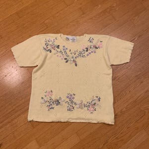 May include: A light yellow short-sleeved sweater with floral embroidery in shades of pink, purple, and blue. The sweater has a scalloped neckline and sleeve edges, and small decorative holes.