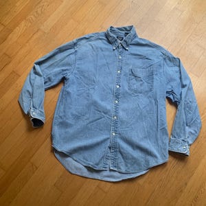 May include: A light blue denim button-down shirt featuring a pointed collar, long sleeves with buttoned cuffs, and a single chest pocket on the left side. The shirt has a central vertical row of white buttons and a Gap brand label at the neck.