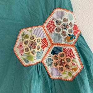 Pu&ograve; includere: Un capo in tessuto turchese con tre toppe a forma esagonale. Ogni toppa presenta un motivo floreale con finiture arancioni. Le toppe sono realizzate con vari tessuti fantasia, tra cui righe, pois e stampe floreali. Il capo ha una trama arricciata.