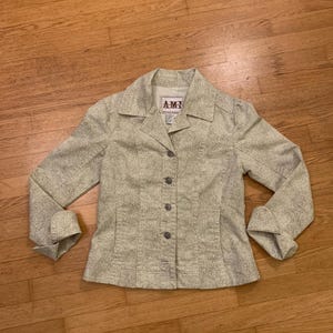 May include: A light green blazer with a subtle dotted pattern. The blazer has a collared neckline, button closure, and cuffed sleeves. The label reads "AMI". The jacket is made of a textured fabric.