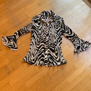May include: A long-sleeve button-down shirt featuring a black, white, and tan zebra-inspired animal print. The garment has a pointed collar, a front button closure, and flared, double-layered ruffled cuffs at the end of each sleeve.