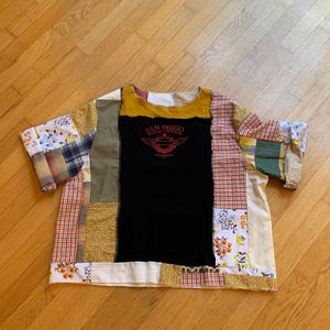 May include: A patchwork t-shirt with a black center panel featuring the text "SAN DIEGO HARLEY-DAVIDSON". The shirt is made of various fabric patterns, including floral, plaid, and solid colors, with a yellow neckline and short sleeves.
