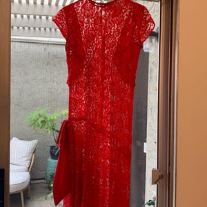 May include: A vibrant red lace dress featuring short sleeves, a round neckline, and a solid red fabric bow detail positioned on the left side of the waist. The garment is constructed from a semi-sheer floral lace pattern and hangs from a white hanger.