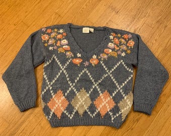May include: A blue argyle sweater with a V-neck and long sleeves. The sweater features a floral design around the neckline and diamond patterns in peach and beige. The sweater is made of a soft knit material.