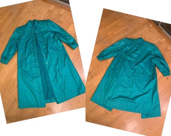 May include: Teal-colored vintage raincoat with a button-up front, long sleeves, and a collared neckline. The coat is made of a shiny, lightweight material and has a classic, A-line silhouette. The coat is displayed on a wooden floor.