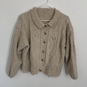 May include: A beige knitted cardigan with a collared neckline, button closure, and a crocheted trim. The cardigan has a textured knit pattern and a relaxed fit.