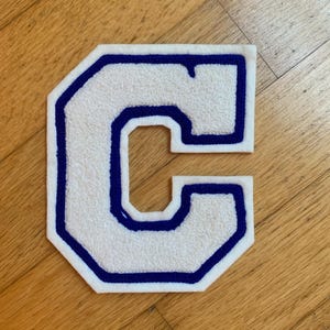 May include: White felt letter "C" with a blue outline. The letter has a textured surface and is set against a wooden background. This patch is suitable for jackets, bags, or other fabric items.