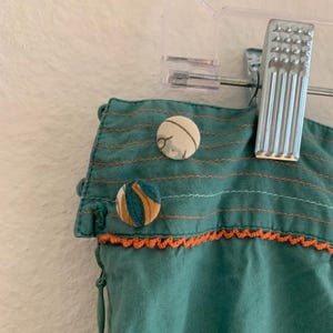 Pu&ograve; includere: Primo piano di tessuto verde acqua con cuciture arancioni e bottoni decorativi. Il tessuto presenta cuciture orizzontali e un bordo all'uncinetto arancione. Sono visibili due bottoni fantasia, uno con strisce blu e gialle e l'altro con un motivo floreale.