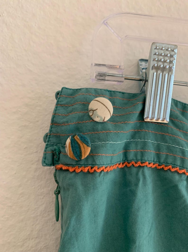 Pu&ograve; includere: Primo piano di tessuto verde acqua con cuciture arancioni e bottoni decorativi. Il tessuto presenta cuciture orizzontali e un bordo all'uncinetto arancione. Sono visibili due bottoni fantasia, uno con strisce blu e gialle e l'altro con un motivo floreale.