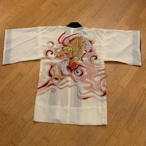 May include: White kimono-style robe with a large, colorful dragon design on the back. The dragon features gold, red, and white scales, with pink and red cloud-like accents. The robe has black trim on the collar and sleeves.