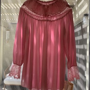Könnte beinhalten: Ein vintage rosa Nachthemd mit langen Ärmeln und einem Rüschenkragen. Das Nachthemd hat Spitzendetails am Kragen und an den Manschetten. Das Kleidungsstück hängt an einem Holzbügel vor einem Fenster.