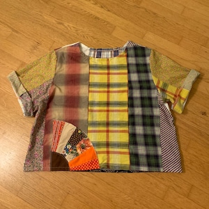May include: A patchwork short-sleeved top with a variety of fabric patterns and colours. The design includes plaid, floral, and striped sections, with a fan-shaped detail at the bottom. The sleeves are rolled up, and the top is laid on a wooden surface.