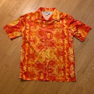 Pode incluir: Camisa de manga curta com um vibrante padrão tie-dye em tons de laranja e amarelo. A camisa tem uma gola clássica e um corte descontraído, perfeito para uso casual. O nome da marca é visível na gola.
