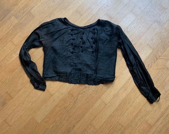 May include: Black long-sleeve blouse with a round neckline and small, dark buttons down the front. The blouse has a gathered waist and cuffs, and is made of a slightly textured fabric. The sleeves are long and loose, and the overall style is vintage-inspired.